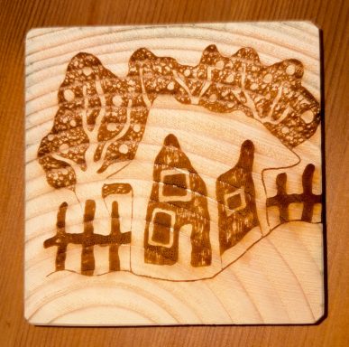 Wooden Coaster featuring a house surrounded by trees and a picket fence. Drawn By Ellie Blundell
